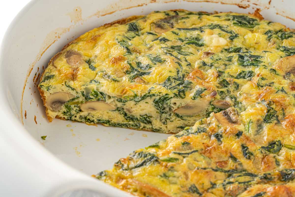 frittata in a casserole dish with a piece cut out, revealing cheesy eggs, spinach and mushrooms