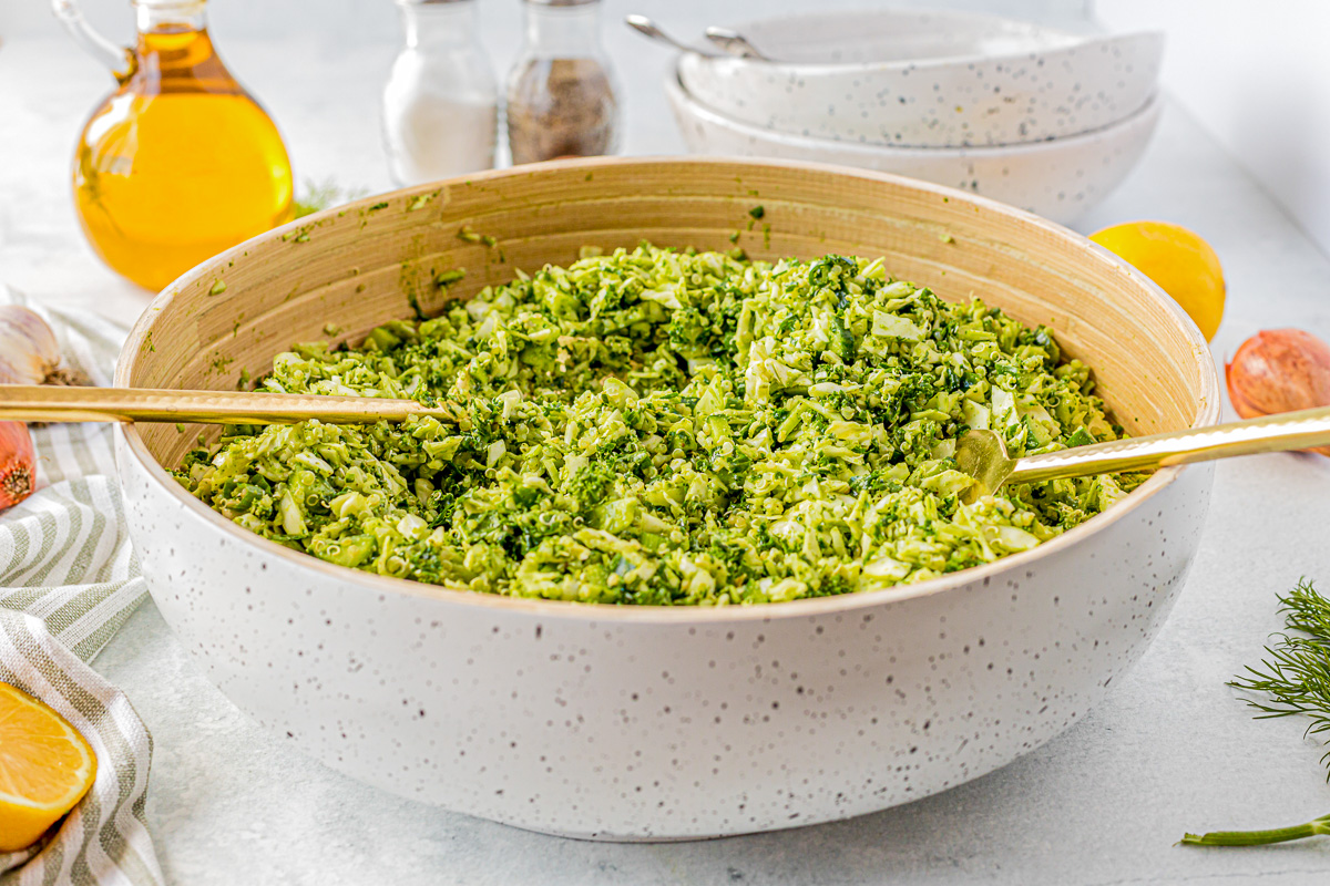 bowl with salad tongs filled with green veggies sitting next to herbs and green veggies