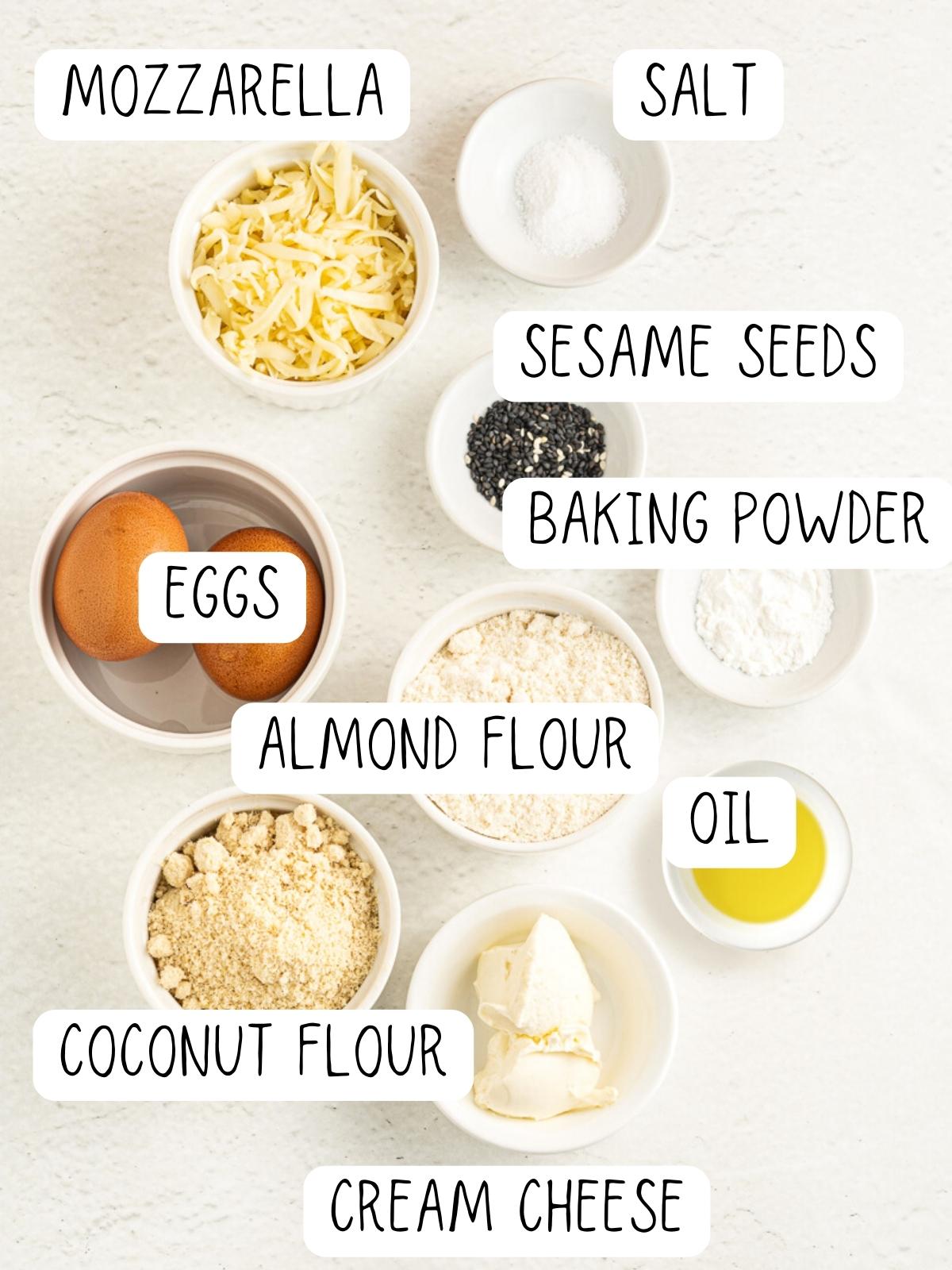 ingredients for keto rolls, including mozzarella cheese, eggs, salts, sesame seeds, baking powder, almond flour, oil, coconut flour and cream cheese